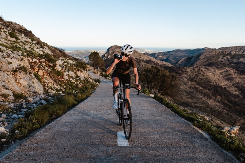 Athlet trinkt einen Schluck aus einer MNSTRY Trinkflasche, während er mit dem Fahrrad in einer bergigen Landschaft unterwegs ist.